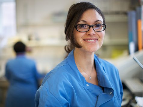 A woman in a blue uniform, at a workbench in a laboratory, smiling