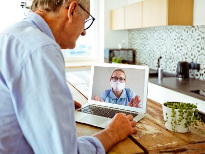 An older man has a video call with a clinician who is wearing a mask