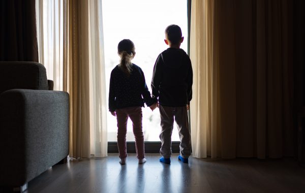 Two children from behind holding hands and looking out of a window