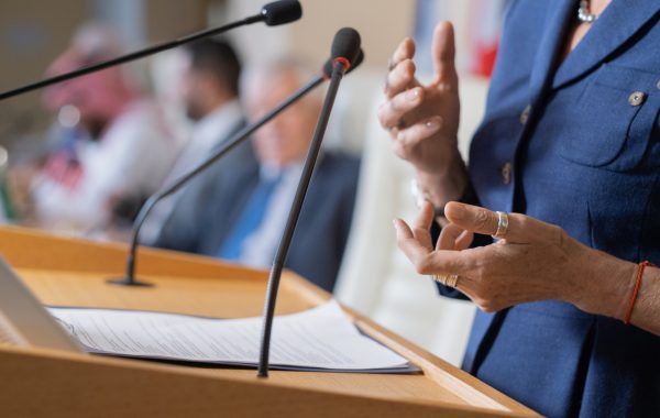 Close-up of unrecognizable female politician in rings standing at rostrum with microphones and clipboard while addressing conference