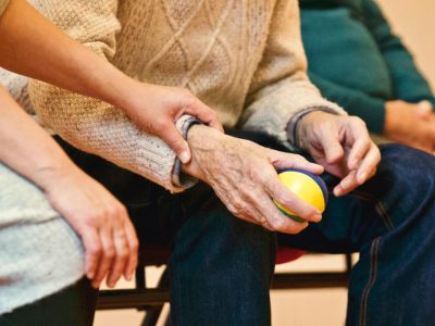 An elderly person is holding a ball in their hand while a healthcare professional guides them during exercise