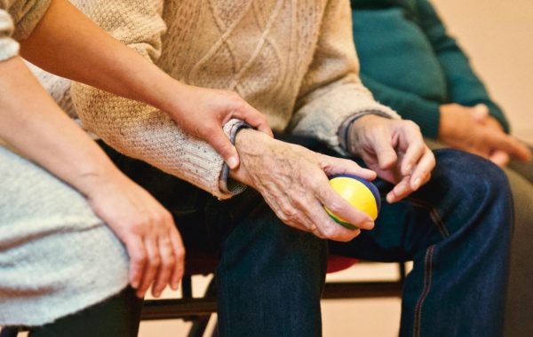 An elderly person is holding a ball in their hand while a healthcare professional guides them during exercise