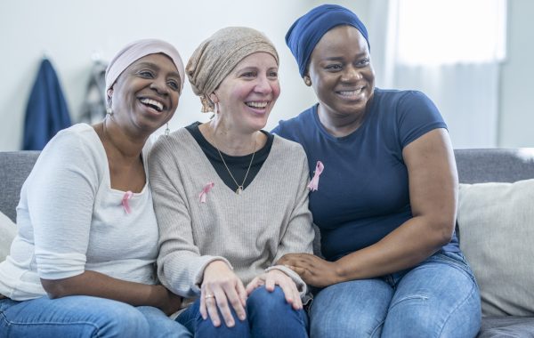A group of three women wearing pink breast cancer awareness ribbons are sitting on a sofa, smiling