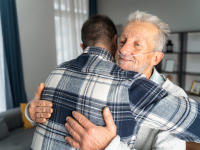 An older man with an oxygen tube embraces a younger man