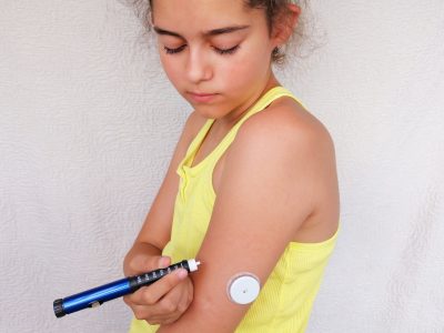 Girl stands and injects insulin from insulin pen in arm. On the arm is also placed white sensor for continuous glucose monitoring in blood