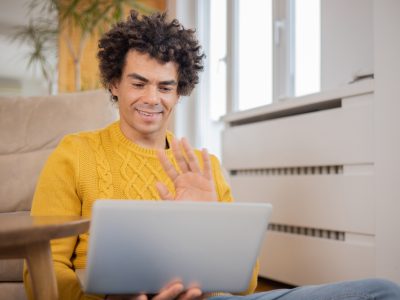 A man wearing a yellow jumper, sitting on the floor of his flat with a laptop on his lap is waving to people he is having an online meeting with