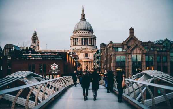 People are walking across a bridge in London with St Paul's cathedral visible in the distance and a sign saying 'City of London'
