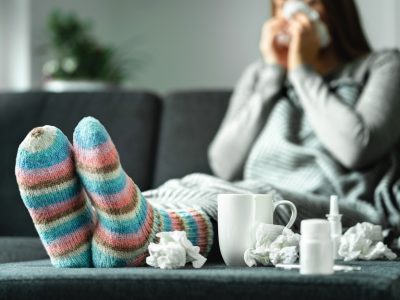 A woman sitting on a sofa with her slippered feet up on a table covered in tissues and cold remedies